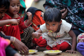 A young hungry child sat right of center in a group of similar young children sat on the ground looks out at the camera hunched over forward while eating from a small bowl in front of him