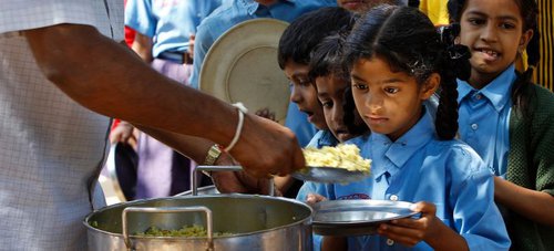 Young children wearing blue collared shirts queue huddled close to receive food served from a large pan